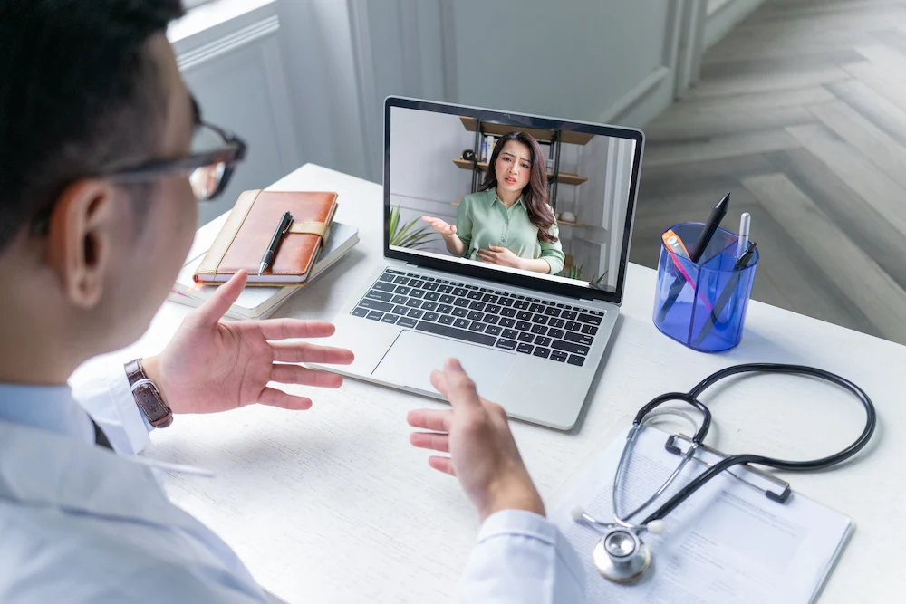 Doctor providing online video consultation to a female patient through a laptop at clinic desk